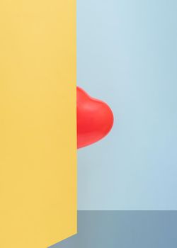 Red heart-shaped balloon hides behind a yellow wall in front of a blue background. Valentines Day. Abstract concept. Upright rectangle layout with copy space.