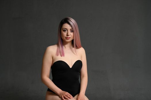 Young woman with dyed hair in a swimsuit sits on a chair on a black background