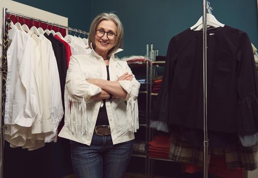 Confident successful elderly European woman seamstress, dressmaker, tailor posing in own fashion design atelier. Clothing designer between hangers with clothing garment in tailoring workshop