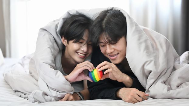 Young gay couple lying under blanket on bed and holding rainbow heart shape. LGBT, pride, relationships and equality concept.