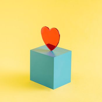 A red heart set on a blue cube isolated on a yellow background. Valentine's Day. Square layout with copy space. Abstract concept. Isometric.
