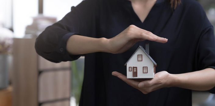 Property insurance and security concept. Protecting gesture of woman and symbol of the house. Wide banner composition with office in background..