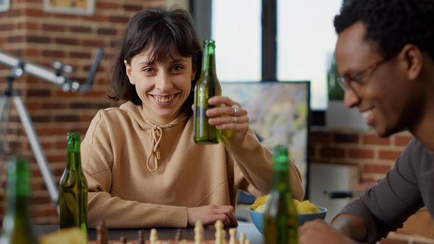 Portrait of happy woman raising beer bottle in front of camera, doing cheers gesture with group of friends at table. Cheerful person enjoying board games play with people for fun acitvity.