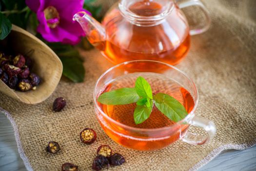 brewed rosehip tea in a glass teapot with rosehip flowers and mint, on a wooden table.