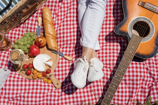 Top view of unrecognizable young woman in white pants outside having picnic, eating and playing guitar. Summer fun and leisure