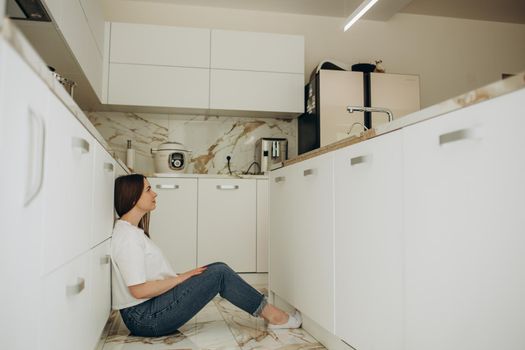 Young woman sitting on the kitchen floor with a paper bag full of fresh groceries with hand on chin thinking about question, pensive expression. Smiling with thoughtful face. Doubt concept