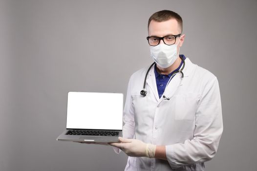 male doctor in a coat, mask, glasses, gloves and with a stethoscope holding a laptop in his hands, isolated screen, studio shooting. looking at the camera