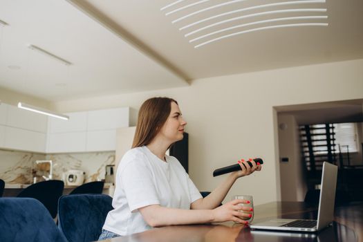 young beautiful woman sitting in the kitchen near the laptop, turns on the TV remote control to the TV