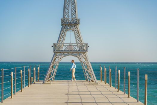 Large model of the Eiffel Tower on the beach. A woman walks along the pier towards the tower, wearing a blue jacket and white jeans