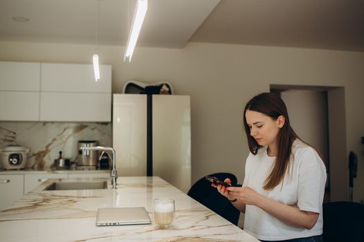 Beautiful woman drinking coffee and using the phone in the kitchen. Young woman enjoying in morning