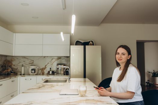 Young woman using smartphone leaning at kitchen table with coffee mug and organizer in a modern home. Smiling woman reading phone message. Brunette happy girl typing a text message.