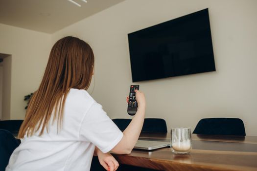 Over the shoulder view of young Asian girl with long black hair sitting on sofa holding tv remote and surfing programs on television. Teenage woman watching TV in the living room.