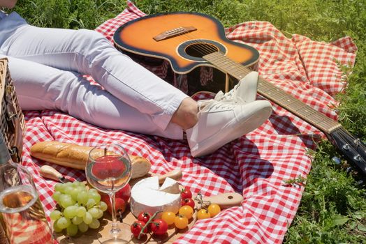 unrecognizable young woman in white pants outside having picnic, eating and playing guitar. Summer fun and leisure