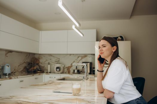 Young attractive smiling Latina woman talking on phone while cooking alone in kitchen. Housewife holds cellphone chatting distracted from healthy vegetarian food preparation. Chores, lifestyle concept.