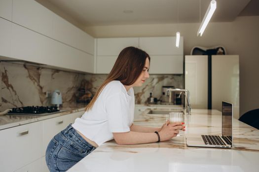 Caucasian female student on video call with friends typing on laptop sipping hot coffee working in kitchen.