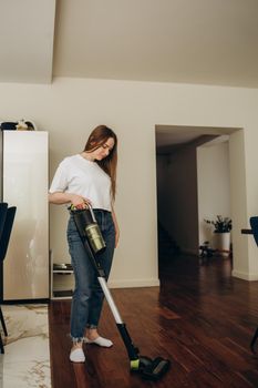 A young Caucasian woman vacuums a kitchen floor using a cordless vertical vacuum cleaner, or electric broom. The maid is cleaning up the hostels cook room. One white female do cleanup of indoors