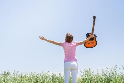 Freedom and happiness. Young woman standing in grass fiel raising her hand to the sky, holding guitar, view from behind