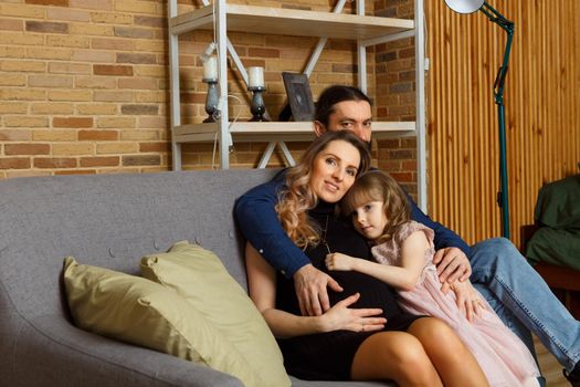 Happy young father, mother and daughter sit on wicker sofa at home. The image of a happy family expecting the second child, studio