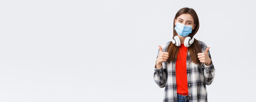 Social distancing, leisure and lifestyle on covid-19 outbreak, coronavirus concept. Cute pleased young caucasian woman in medical mask, take-off headphones to say yes or good, make thumb-up sign.