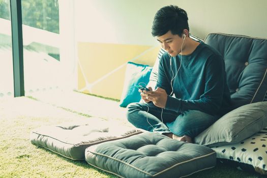 asian male teenager man listening to music from smart mobile phone while sitting on floor at home