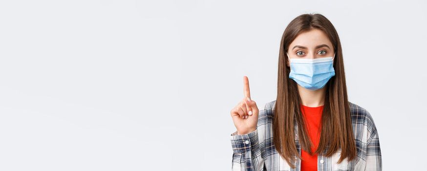 Coronavirus outbreak, leisure on quarantine, social distancing and emotions concept. Close-up of cute young woman in medical mask explain smth, show number one or point up.