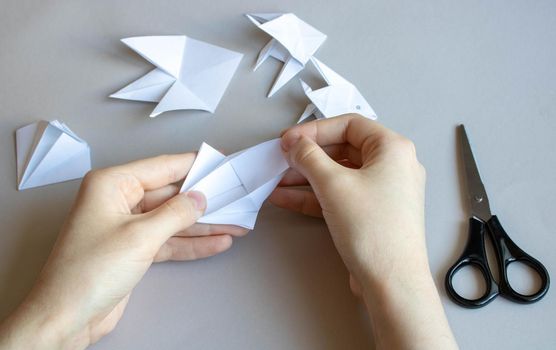 Hands make white paper fish on a gray table.There are scissors next to it.