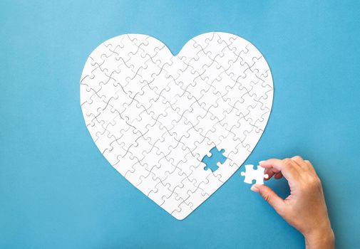 White puzzle in heart shape. Hand with white details of puzzle on blue background.