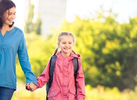 Little girl going to school with mother holding hand and smiling on sunny nature background