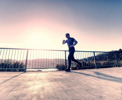 Tall slim man run on the lake terrace against the backdrop of a beautiful sunrise. Shore of mountain lake with dark forest hills