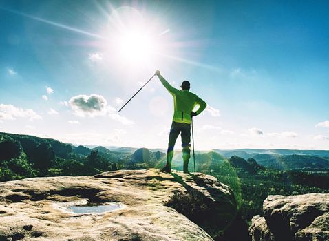 Funny slim blond tourist guy with backpack holds trekking stick on rocky top.  Foggy mountain range panorama with man silhouette.