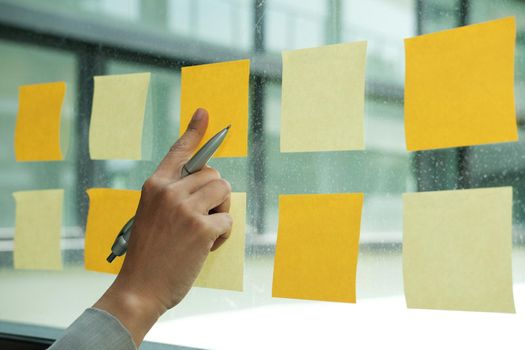 hand pointing at adhesive notes on glass wall at workplace. Sticky note paper reminder schedule for creative idea & business brainstorming