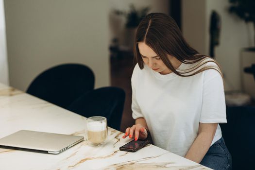 Young woman using smartphone leaning at kitchen table with coffee mug and organizer in a modern home. Smiling woman reading phone message. Brunette happy girl typing a text message