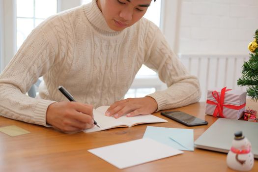 man wearing cream sweater writing note on notebook at home during christmas new year holiday celebration.