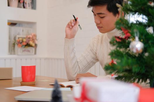 man wearing cream sweater writing note on notebook at home during christmas new year holiday celebration.