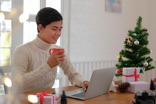 man wearing cream sweater holding coffee cup & using computer at home during christmas. xmas new year celebration