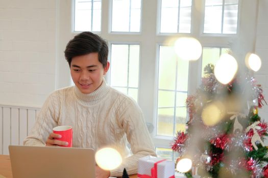 man wearing cream sweater holding coffee cup & using computer at home during christmas. xmas new year celebration