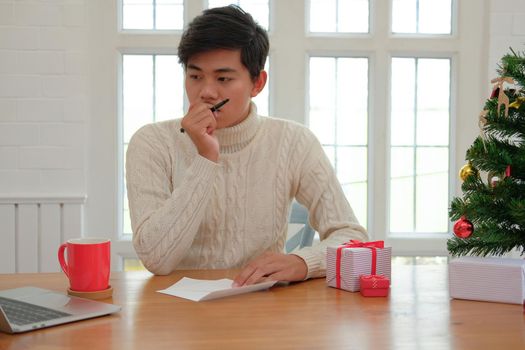 man wearing cream sweater thinking what to write on christmas letter greeting card holiday wishes with xmas decoration.
