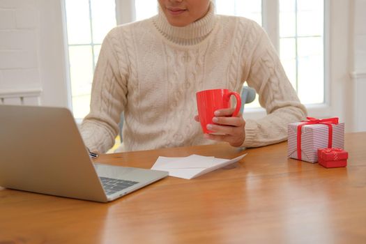 man wearing cream sweater holding coffee cup & using computer at home during christmas. xmas new year celebration