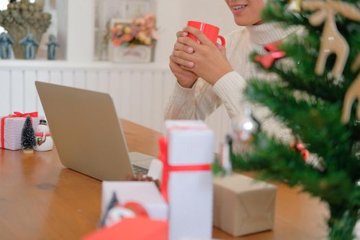 man wearing cream sweater holding coffee cup & using computer at home during christmas. xmas new year celebration