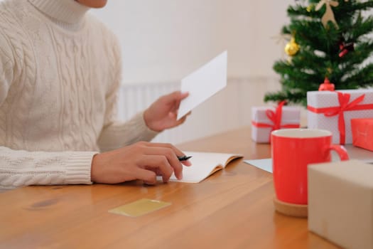 man wearing cream sweater writing christmas letter greeting card holiday wishes with xmas decoration.