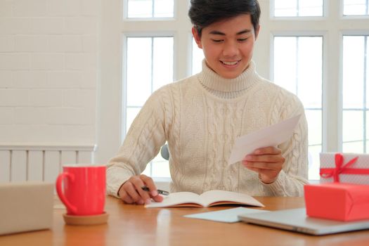 man wearing cream sweater writing christmas letter greeting card holiday wishes. xmas celebration.