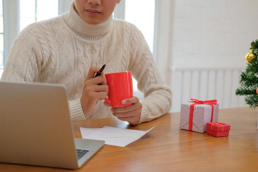 man wearing cream sweater holding coffee cup & using computer at home during christmas. xmas new year celebration