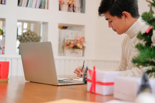 man wearing cream sweater writing christmas letter greeting card holiday wishes with xmas decoration.