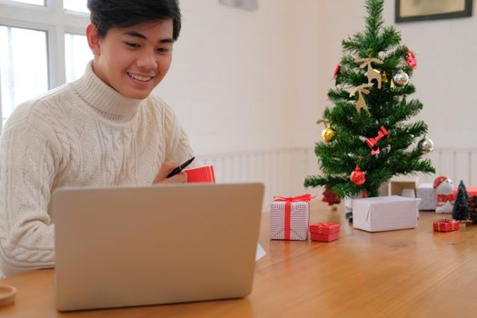 man wearing cream sweater holding coffee cup & using computer at home during christmas. xmas new year celebration