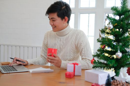man wearing cream sweater holding coffee cup & using computer at home during christmas. xmas new year celebration