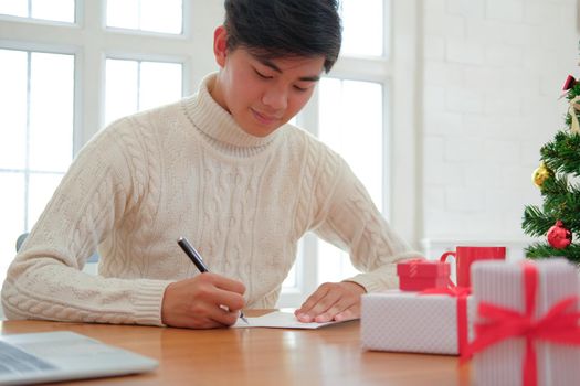 man wearing cream sweater writing christmas letter greeting card holiday wishes with xmas decoration.
