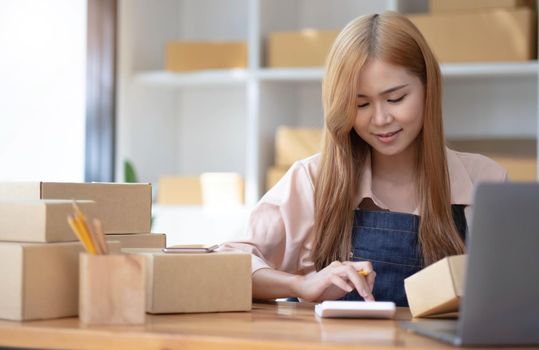 Startup small business SME. Beautiful asian entrepreneur using calculator, check online order and prepare to packing for customer. Successful SME business online concept..