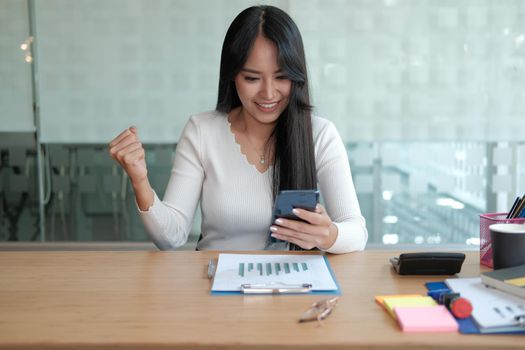 businesswoman holding mobile phone feeling happy glad showing yes gesture at office. young asian freelance woman female entrepreneur using smartphone. business concept
