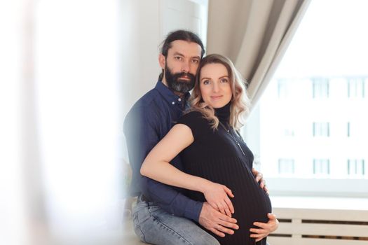 Man embracing pregnant partner in kitchen. Adult family pregnancy concept. Future parents in home outfit embrace standing in the kitchen, looking at each other, kissing. Healthy Lifestyle.