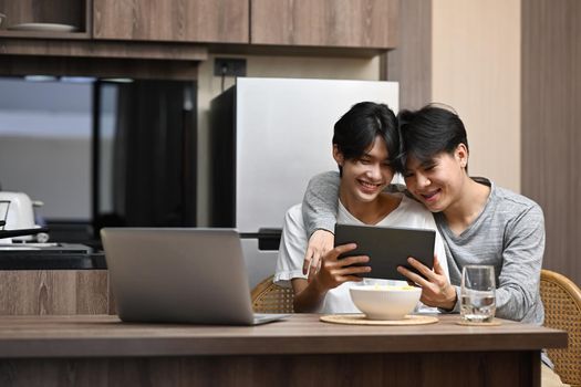 Happy gay couple embracing and surfing internet with digital tablet together at kitchen table. LGBT, pride, relationships and equality concept.